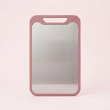 A pink cutting board with a stainless steel surface and integrated handle stands against a pale pink background.