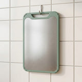 A rectangular kitchen cutting board with a brushed stainless steel center and a sage green frame, hanging on a white tiled wall.