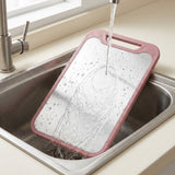 A pink-framed stainless steel cutting board rests diagonally in a kitchen sink beneath running water.