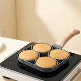 Four-well gray speckled egg frying pan with a wood-grain handle cooking four golden pancakes on a black countertop burner.