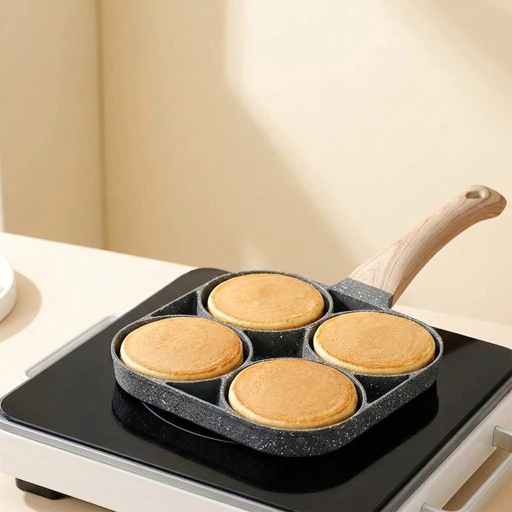 Four-well gray speckled egg frying pan with a wood-grain handle cooking four golden pancakes on a black countertop burner.