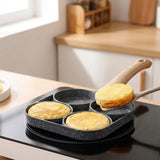 A dark speckled four-well egg frying pan sits on a black kitchen cooktop as a spatula lifts one of three golden pancakes.