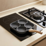 A gray speckled four-well egg frying pan with a wood-grain handle rests on a black glass cooktop beside a gas burner.