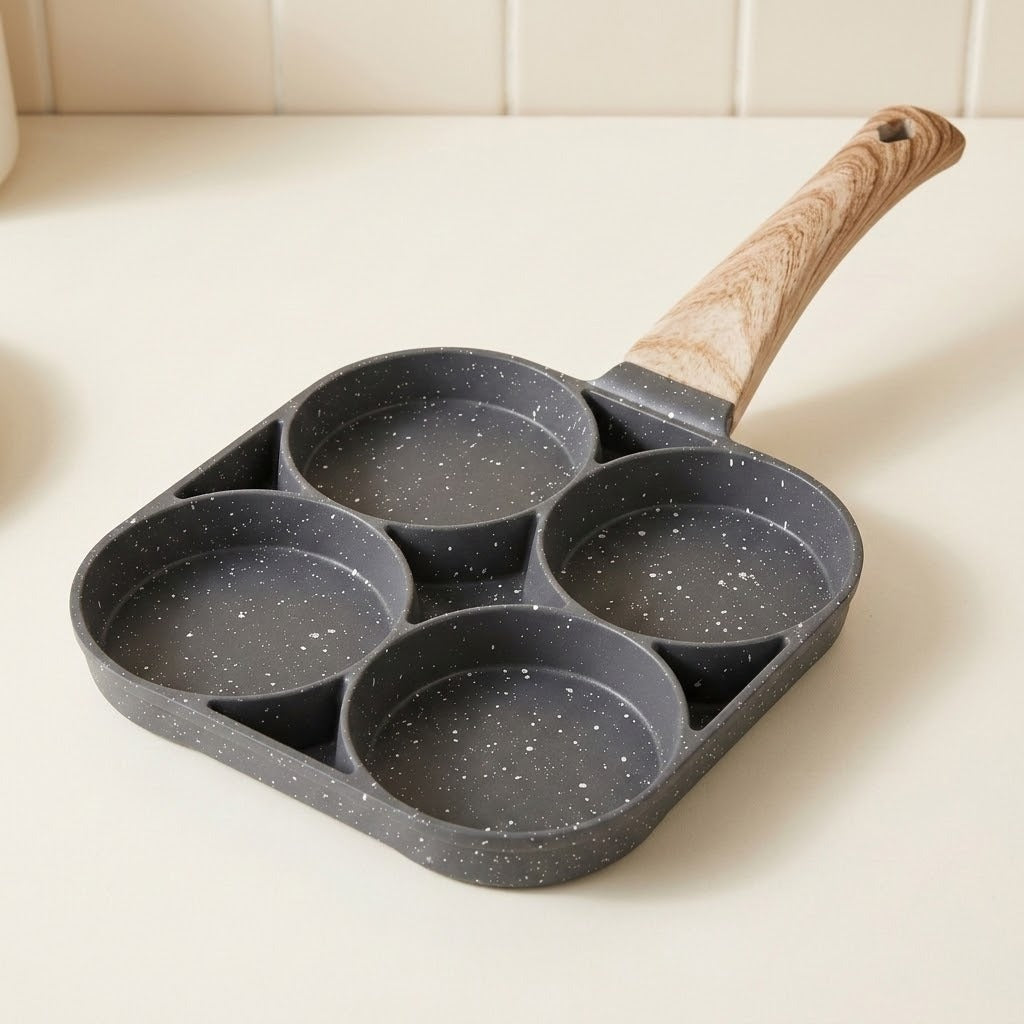 Empty dark gray speckled egg frying pan with four round wells and a wood-grain handle on a cream kitchen countertop.