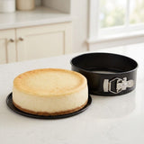 A black springform cake pan with a silver clasp, shown empty and standing beside a freshly baked cheesecake on a white countertop in a bright kitchen.