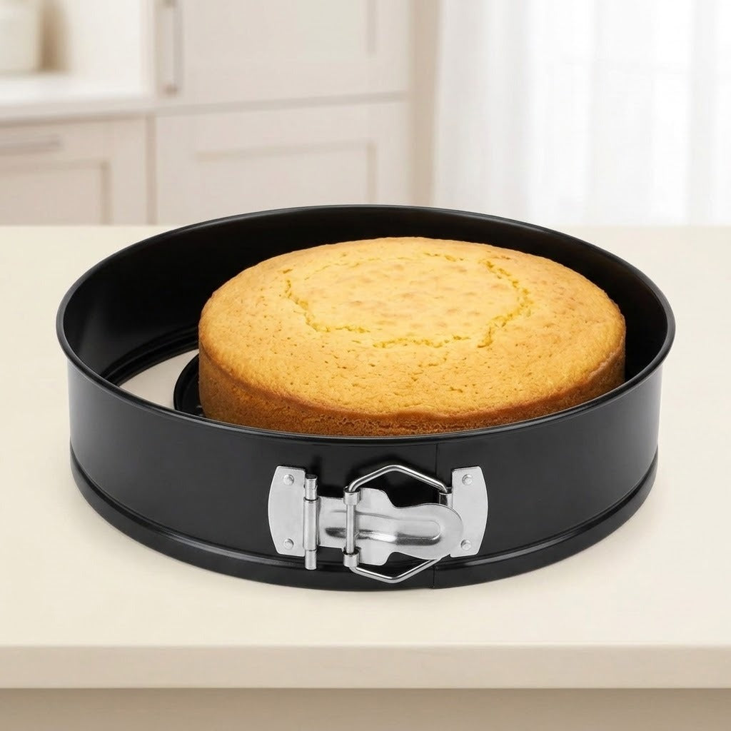 A round black springform cake pan with a silver latch, holding a golden-brown round cake on a light countertop in a softly blurred kitchen setting.
