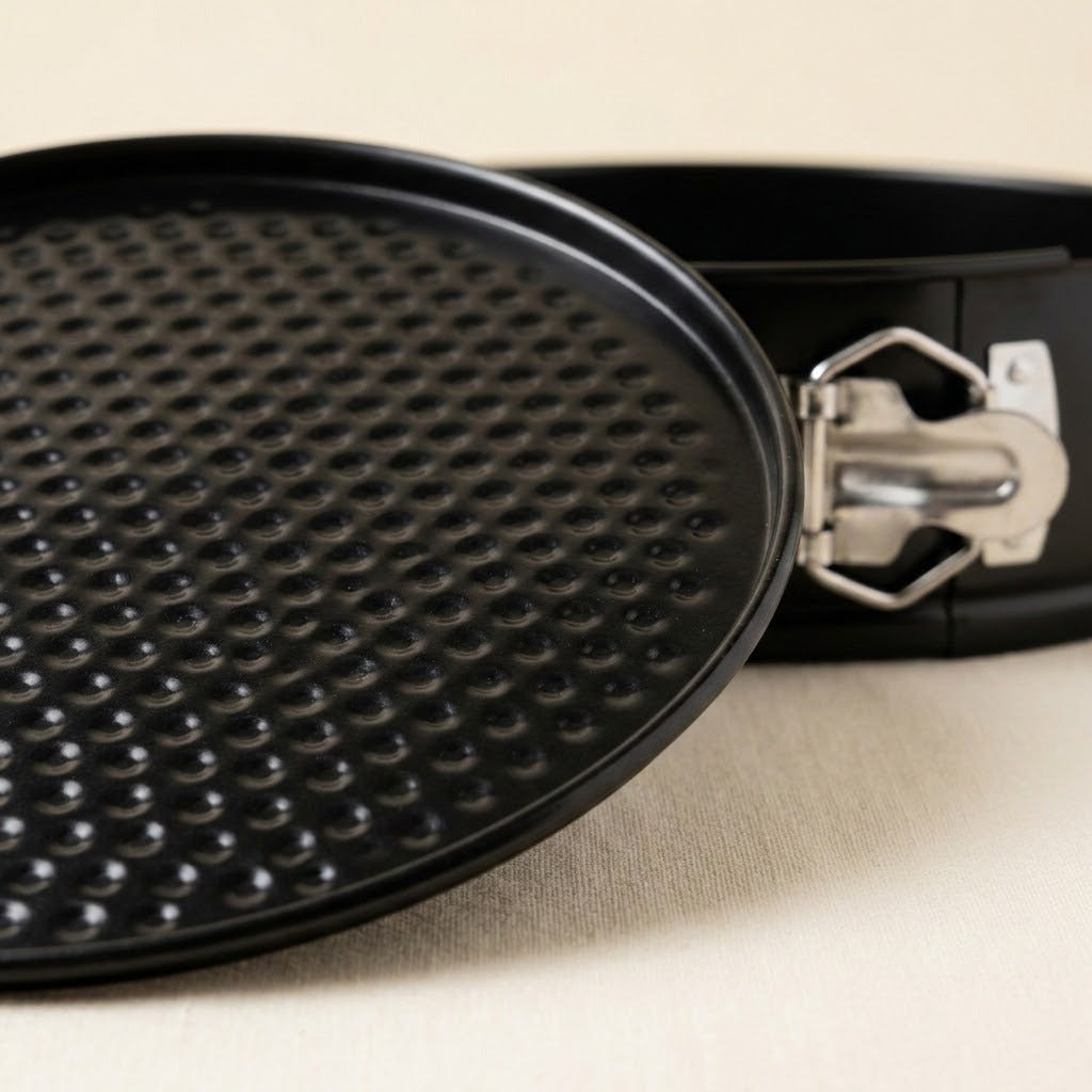 Close-up of a black springform pan showing the textured non-stick inner dish and the metal clasp on a light beige fabric background.