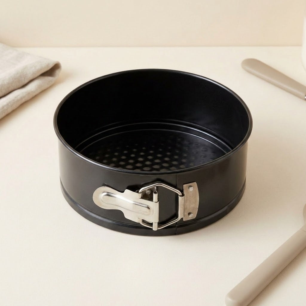A black non-stick springform cake pan with a silver metal clasp, shown on a light beige tabletop with a folded cloth and utensils in the background.