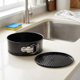 A black springform cake pan with its lid placed beside it on a light beige kitchen countertop near a sink.