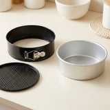 A round springform cake pan set on a light beige countertop, including a black ring with a silver clasp, a silver metal cake pan, and a black non-stick liner.