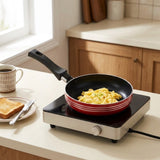 A red and black non-stick frying pan with a black handle sits on a white electric burner on a kitchen counter, with scrambled eggs cooking inside. A cup of coffee, a slice of toast, and a knife are visible on the left, and a kitchen background is in soft focus.