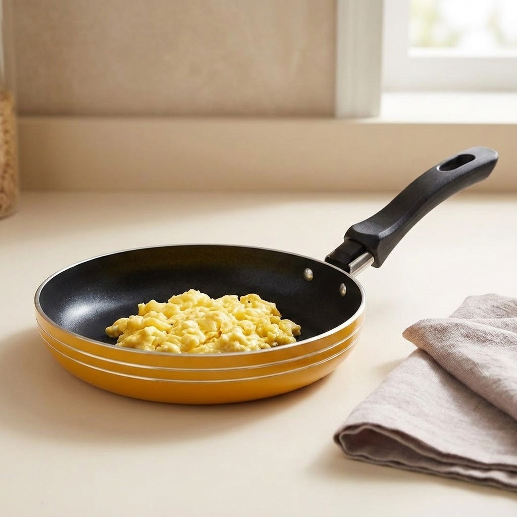 A yellow nonstick frying pan with a black handle sits on a beige countertop, containing scrambled eggs. A folded beige cloth is on the right, and a window and wall are in the background.
