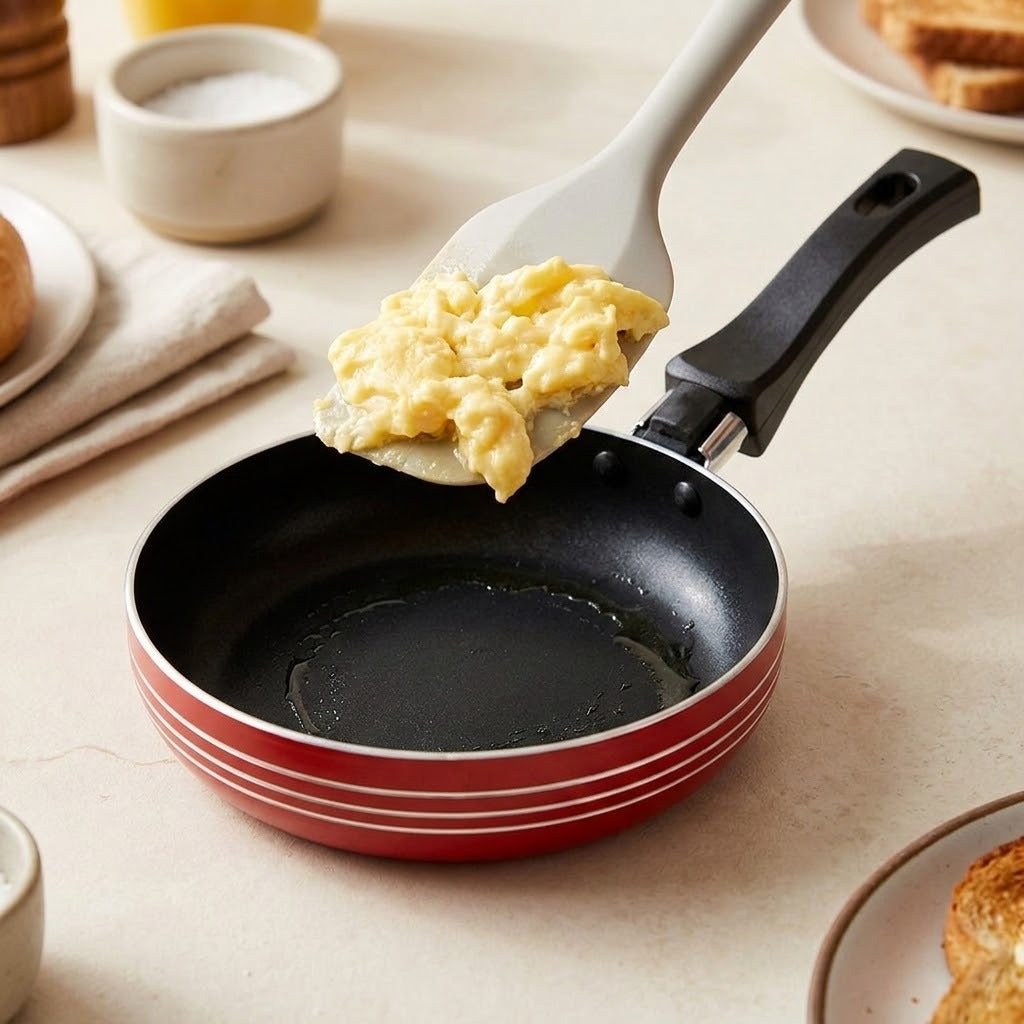 A small red and black frying pan with a white spatula holding scrambled eggs, set on a light beige tabletop with breakfast items in the background.