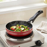 A red and black non-stick frying pan with a black handle sits on a portable electric burner in a kitchen setting, cooking broccoli, zucchini, and red peppers.