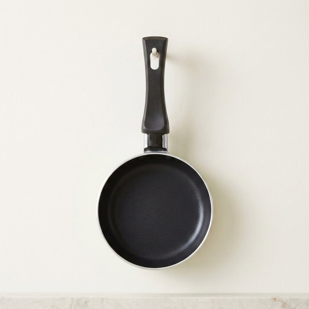 A black non-stick frying pan with a matching black handle, hanging against a plain off-white wall.