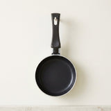 A black non-stick frying pan with a matching black handle, hanging against a plain off-white wall.