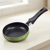 A non-stick frying pan with a black interior and a green exterior, featuring a black handle, resting on a light beige countertop near a window.