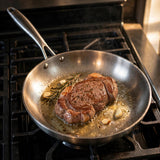 Silver metal frying pan holds a browned steak with garlic and rosemary in bubbling fat on a gas stovetop.