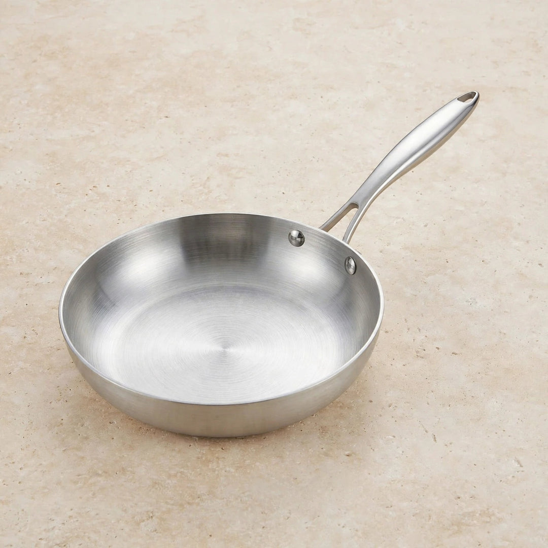 Empty silver metal frying pan with a long riveted handle rests on a beige speckled countertop.