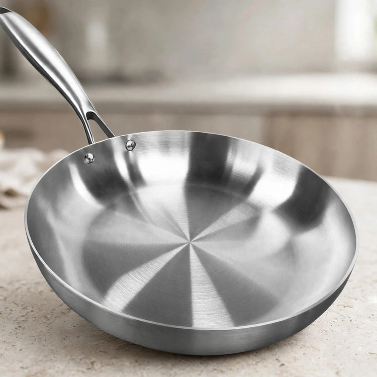 Close-up of a silver metal frying pan with a brushed circular interior and riveted handle on a light kitchen countertop.