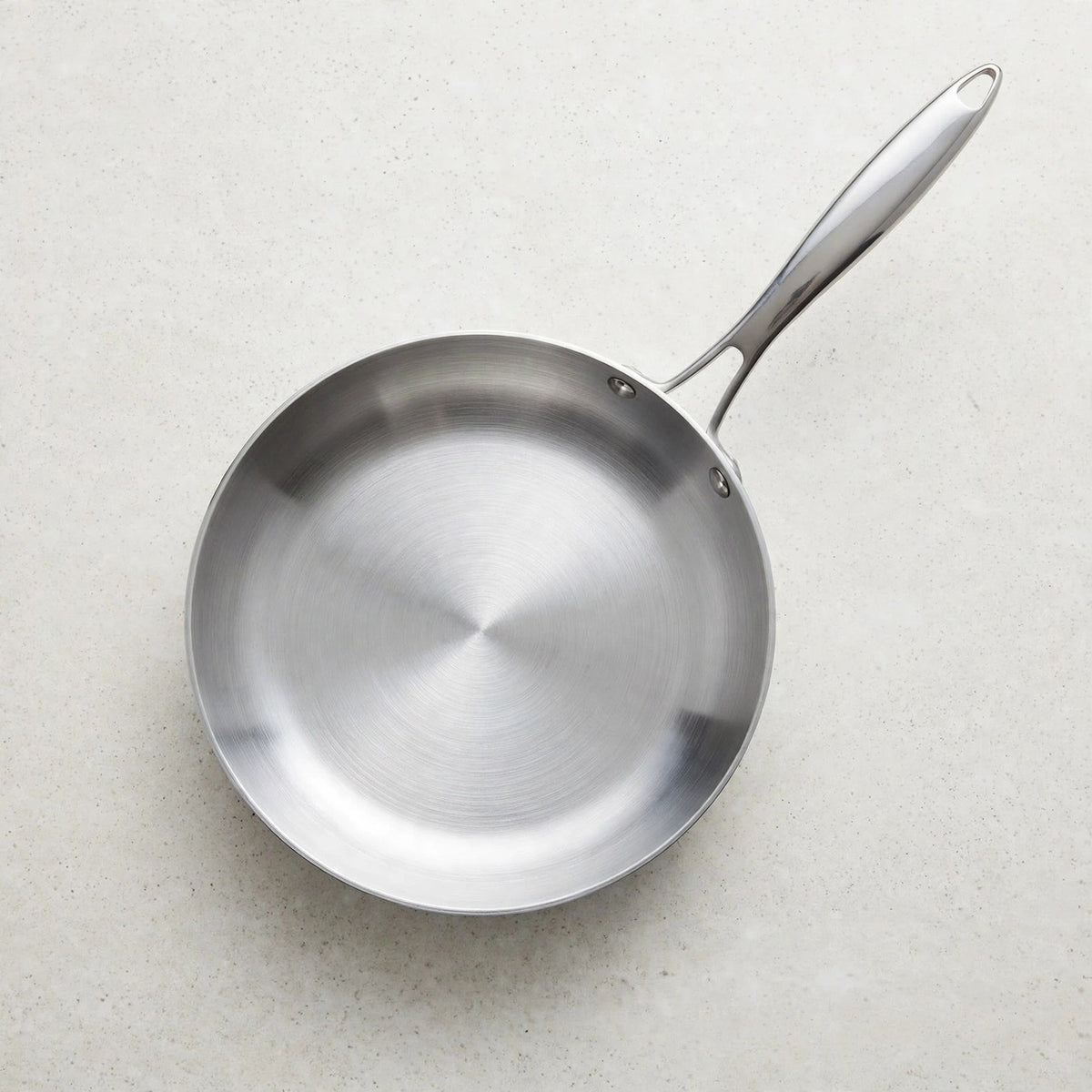 Empty silver metal frying pan viewed from above on a pale speckled countertop, with its handle pointing upper right.