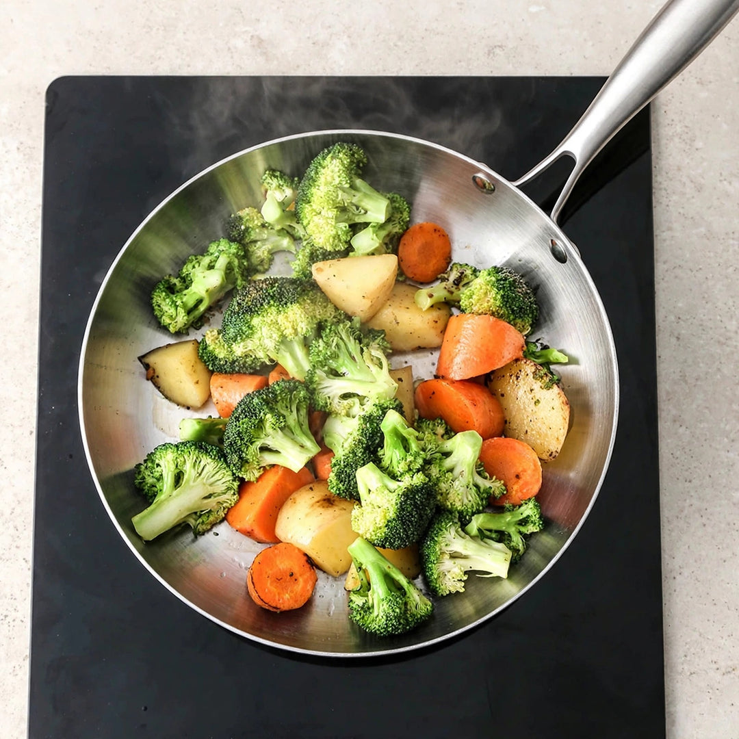 Overhead view of a silver metal frying pan containing steaming broccoli, carrots, and potatoes on a black glass cooktop.