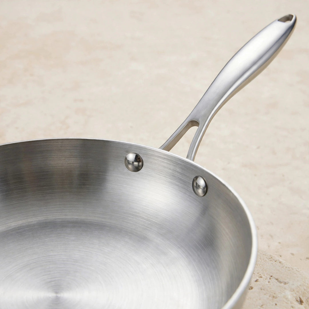 Close-up of a silver metal frying pan showing its brushed interior, two handle rivets, and long handle against a beige background.