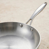 Close-up of a silver metal frying pan showing its brushed interior, two handle rivets, and long handle against a beige background.