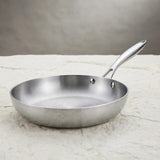 A silver metal frying pan with a long riveted handle rests on a pale stone countertop against a grey background.
