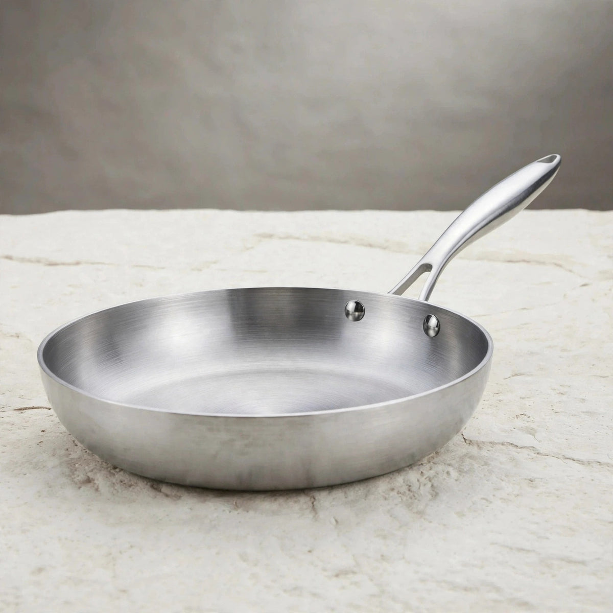 A silver metal frying pan with a long riveted handle rests on a pale stone countertop against a grey background.