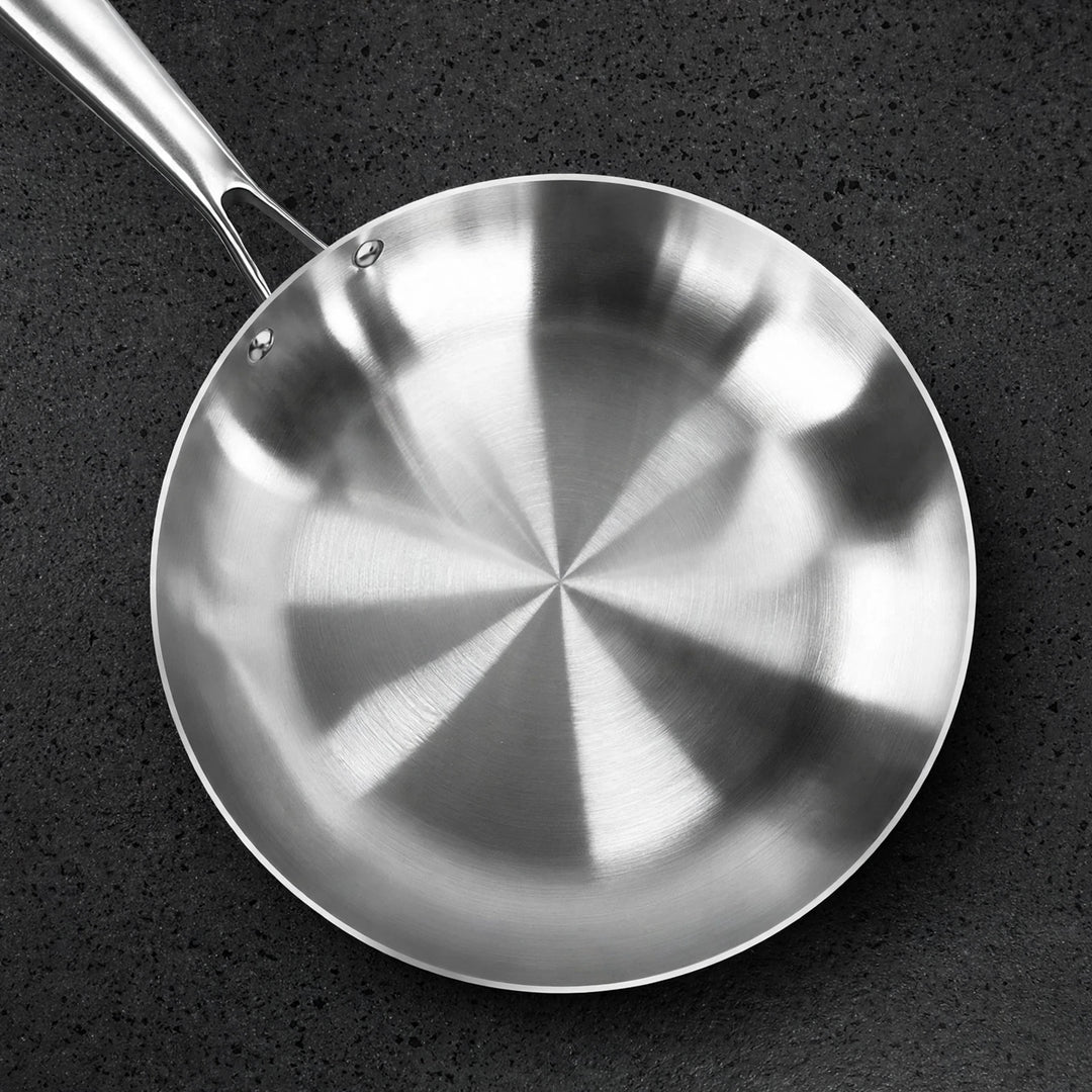 Overhead view of an empty silver metal frying pan with a reflective interior on a dark textured surface.