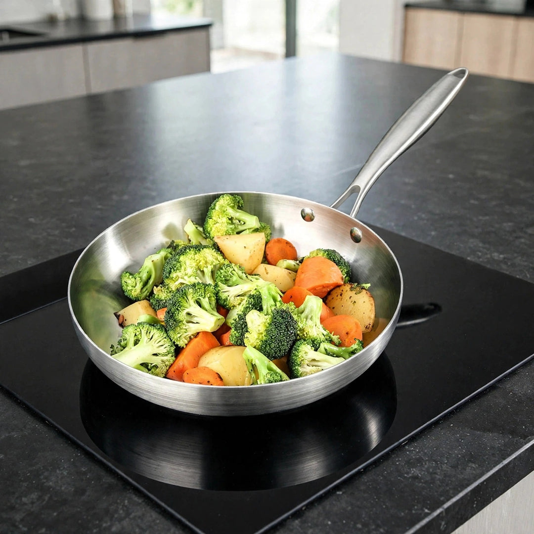 Silver metal frying pan filled with broccoli, carrots, and potatoes sits on a black glass cooktop in a bright kitchen.