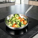 Silver metal frying pan filled with broccoli, carrots, and potatoes sits on a black glass cooktop in a bright kitchen.