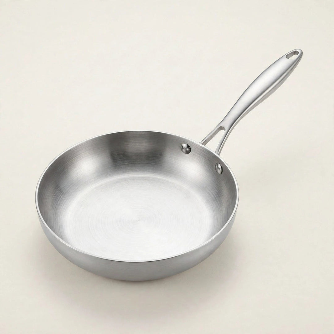 A silver metal frying pan rests on a cream background, showing its round interior and long handle with a hanging hole.