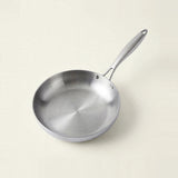 A silver metal frying pan sits on a cream background, with its brushed interior visible and handle angled toward the upper right.