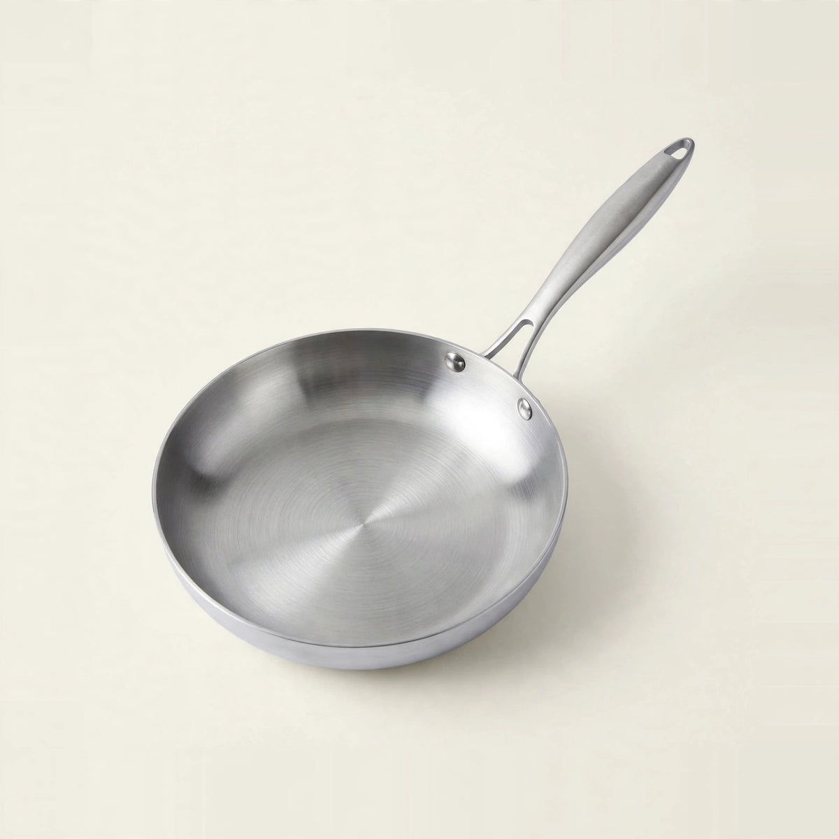 A silver metal frying pan sits on a cream background, with its brushed interior visible and handle angled toward the upper right.