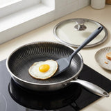 A stainless steel frying pan with a dark gray non-stick interior sits on a black induction cooktop, cooking a sunny-side-up egg. A matching glass lid with a silver knob rests behind it on a kitchen counter.