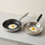 Two stainless steel frying pans with textured non-stick interiors sit on a light gray countertop, each holding a sunny-side-up egg.
