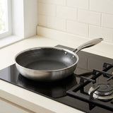 A stainless steel frying pan with a textured dark non-stick interior sits on a black gas stovetop in a kitchen setting with cream tile backsplash.