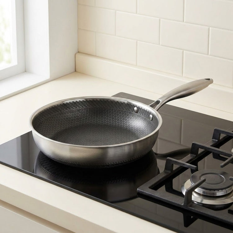 A stainless steel frying pan with a textured dark non-stick interior sits on a black gas stovetop in a kitchen setting with cream tile backsplash.
