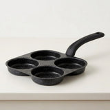 A black speckled non-stick pan with four round cavities and a single curved handle, photographed on a clean white surface against a white background.
