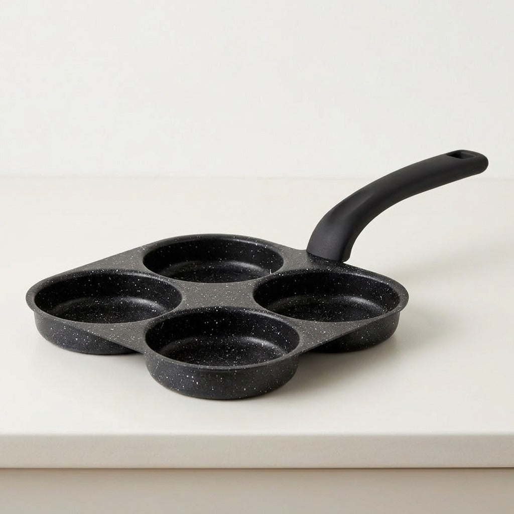 A black speckled non-stick pan with four round cavities and a single curved handle, photographed on a clean white surface against a white background.
