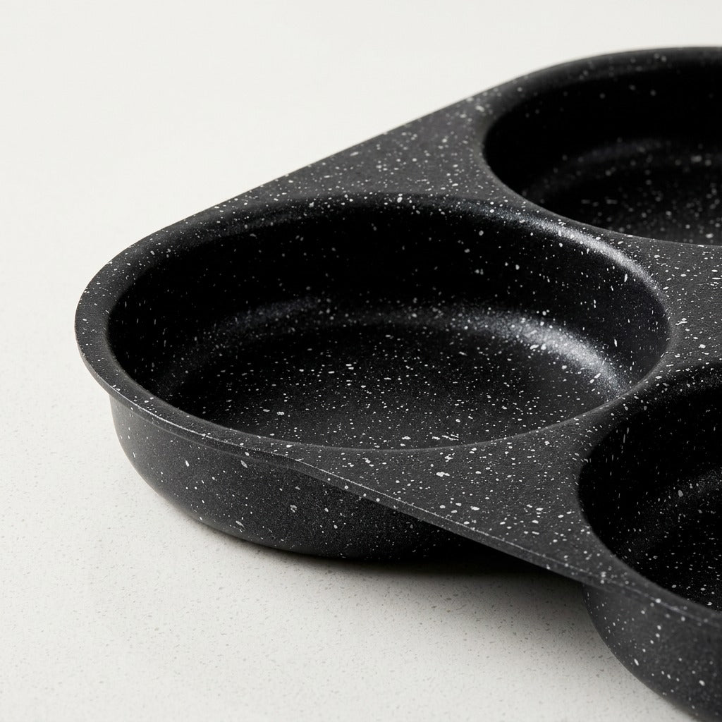 A close-up of a black speckled non-stick three-compartment pan or mold, showing the textured interior of one compartment and the handle on a light gray background.