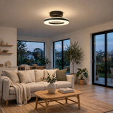 A modern ceiling light fixture with a black circular frame and a glowing white ring, mounted on the ceiling of a contemporary living room with a beige sofa, wooden coffee table, and large windows.