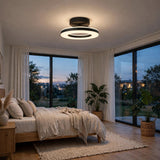 A modern circular ceiling light fixture with a black outer ring and white illuminated inner rings, mounted on the ceiling of a contemporary bedroom with large windows, plants, and a wooden bed.