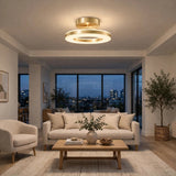 A modern circular ceiling light fixture with a brushed gold finish and warm white glow, installed in a bright contemporary living room with a sofa, coffee table, and large windows overlooking a city skyline at dusk.
