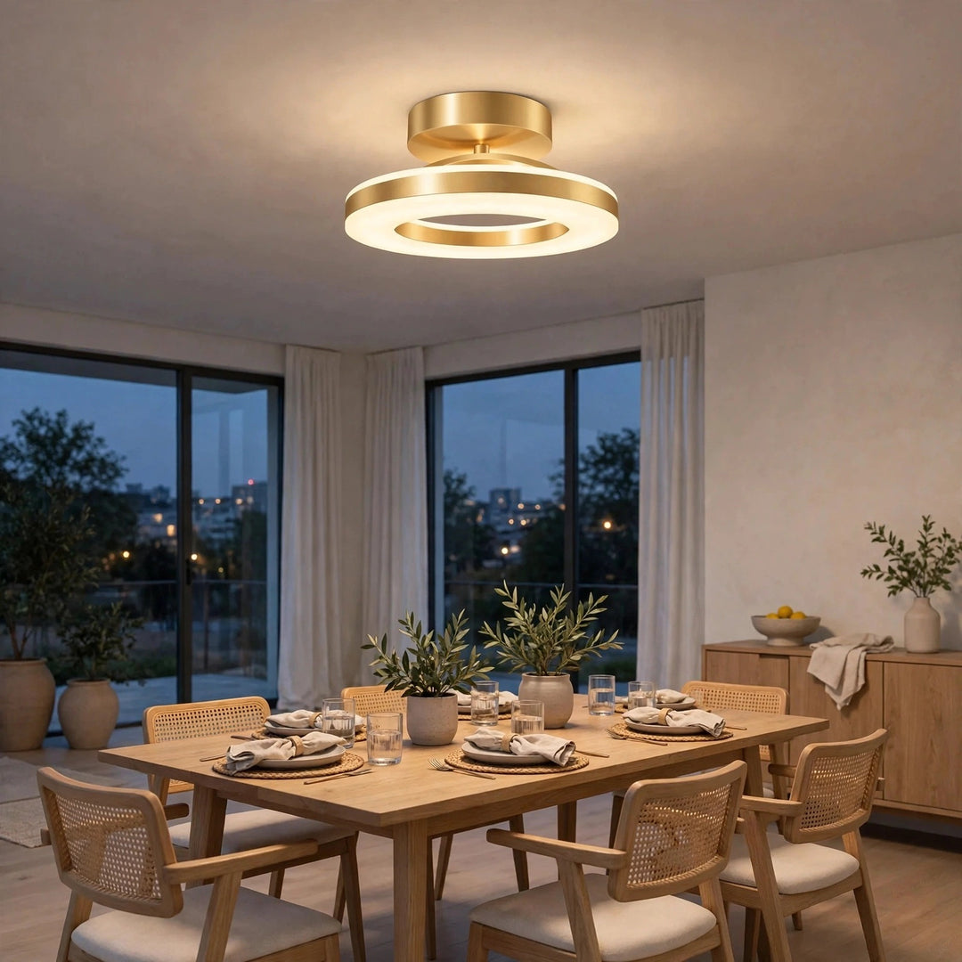 A modern double-ring ceiling light fixture with a brushed gold finish is illuminated in a dining room setting. The room features a wooden dining table, woven-back chairs, and large windows looking out onto a twilight outdoor scene.