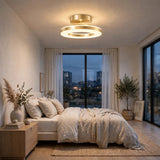 A modern circular ceiling light fixture with a brushed gold finish is illuminated in a beautifully styled bedroom with neutral beige bedding, a wooden nightstand, and large windows overlooking a city skyline at dusk.
