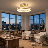 A modern double-ring LED ceiling light with a brushed gold finish is illuminated in a bright, contemporary living room with large windows overlooking a city skyline at dusk.