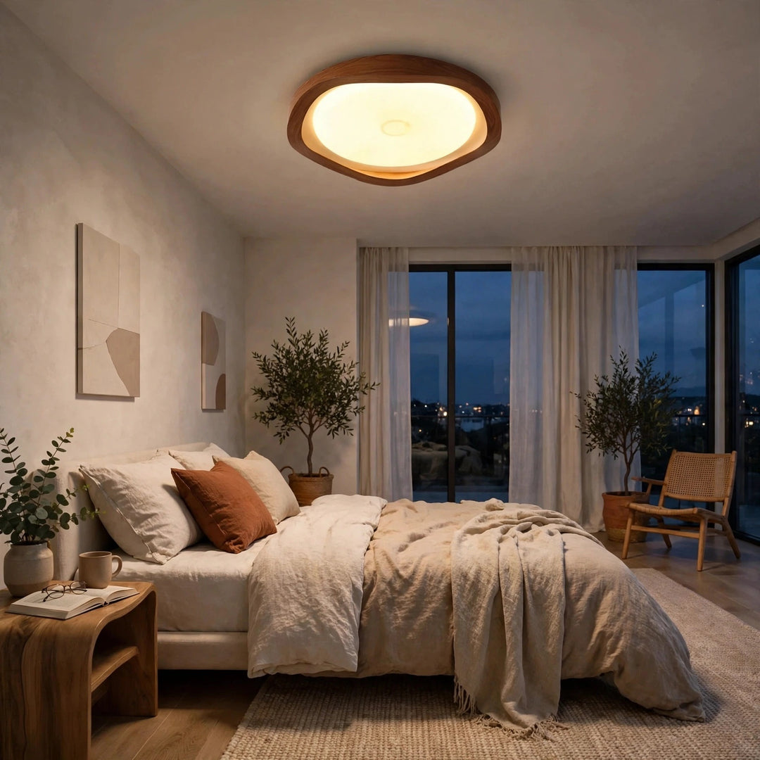 A modern bedroom interior with a bed dressed in neutral beige bedding, a wooden nightstand, potted plants, and a large oval ceiling light glowing above. Large windows with sheer curtains look out onto a dark evening sky.